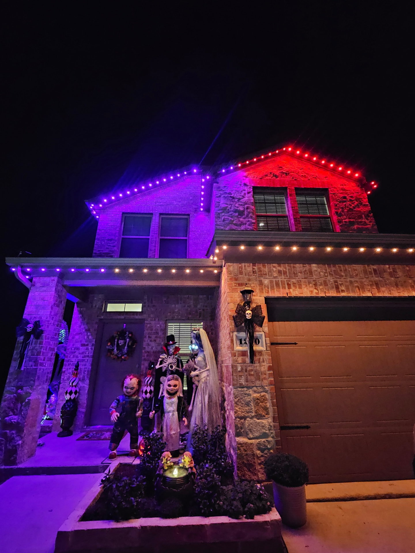 Brick-and-stone home with red and purple permanent lighting along the front gables and Halloween decorations beside the entry