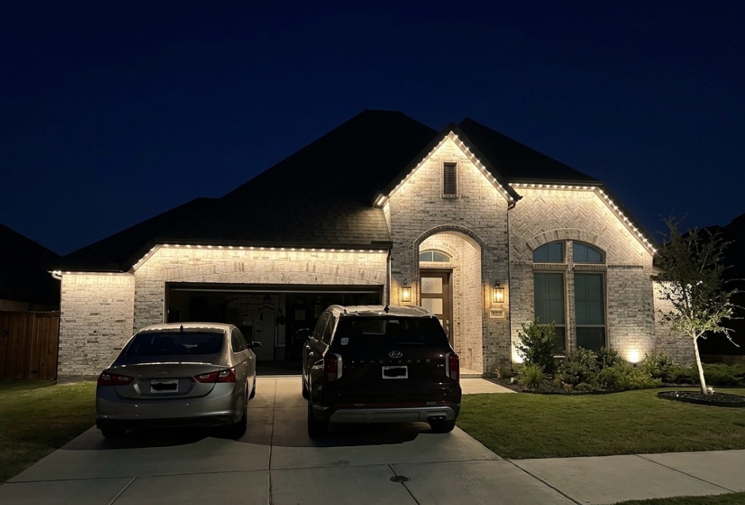 The same light-brick home at night with warm-white permanent lighting mounted behind the fascia around the front roofline